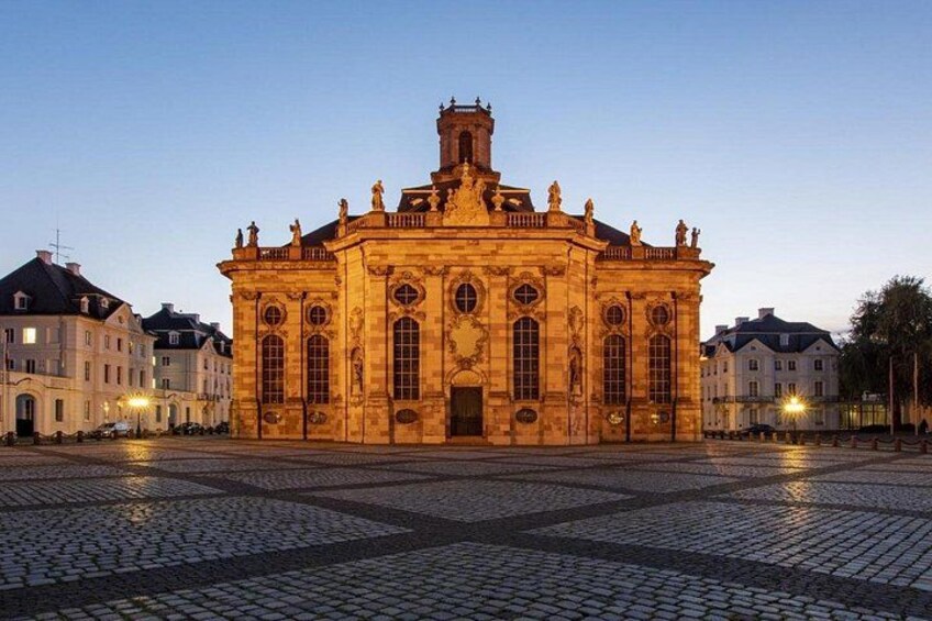 Un bâtiment historique à l’architecture riche et ornée, une place pavée et des bâtiments environnants. 1