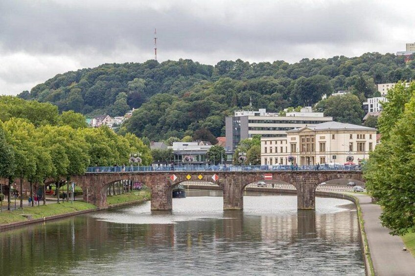 Une rivière traversée par un pont de pierre, un paysage urbain avec des bâtiments et une colline parsemée d’arbres en arrière-plan. 3