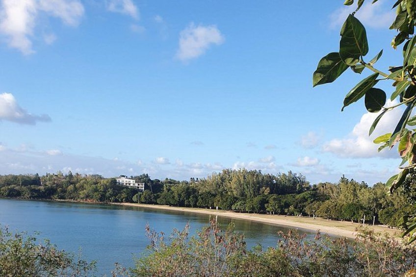 A calm lake with a sandy beach, surrounded by dense greenery and a few buildings in the distance. 5