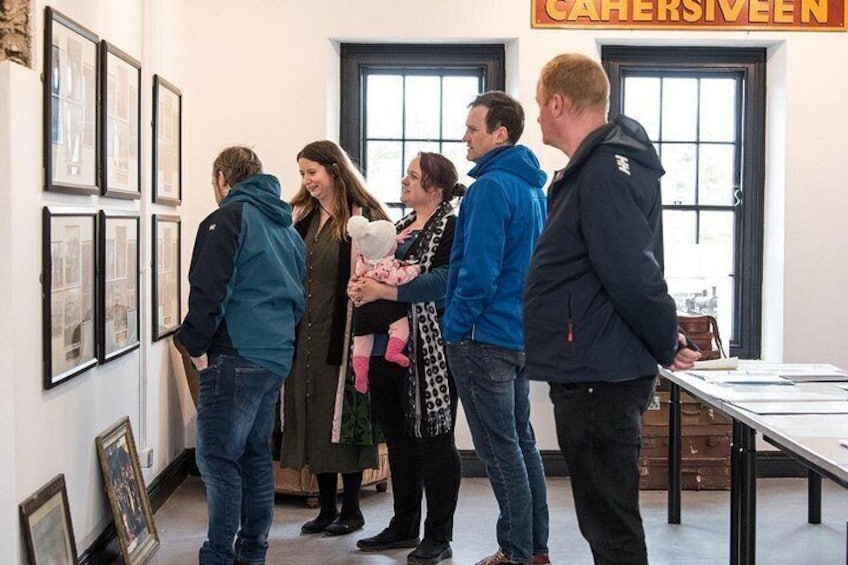 A group of people in a room with framed pictures on the wall and a sign that reads 'CAHERSIVEEN'. 1