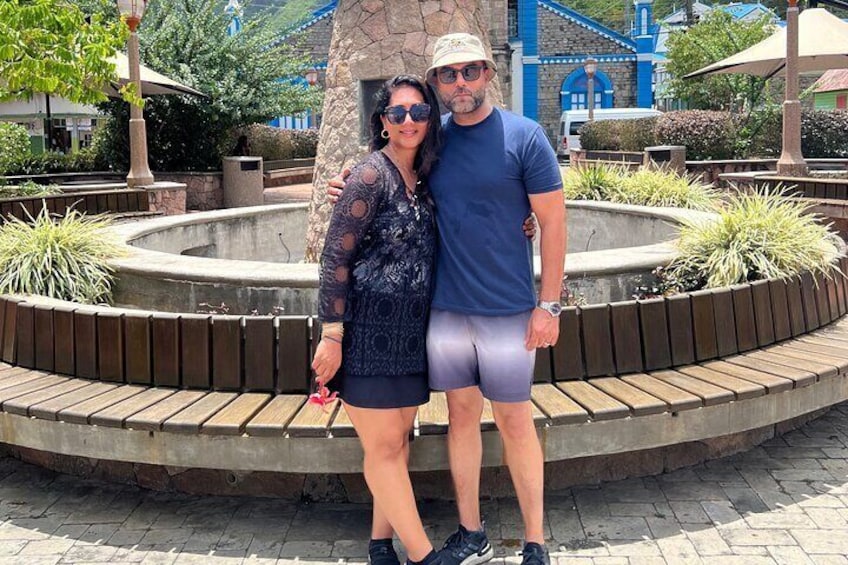 Deux personnes se tiennent devant une fontaine dont le décor est constitué d’un motif circulaire en pierre, et qui est entourée de plantes. 5
