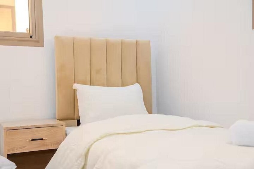 A bed with a beige headboard, white bedding, and a wooden nightstand with a drawer. 5