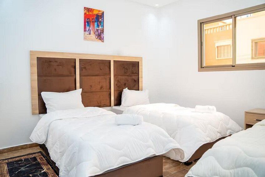 A hotel room with two beds, a wooden headboard, white bedding, a small bedside table, and a painting on the wall. 2