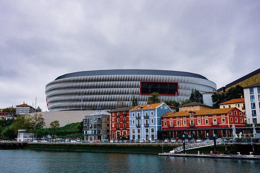 Ein modernes Stadion mit geschwungener Architektur, umgeben von farbenfrohen historischen Gebäuden und einem Gewässer. 3