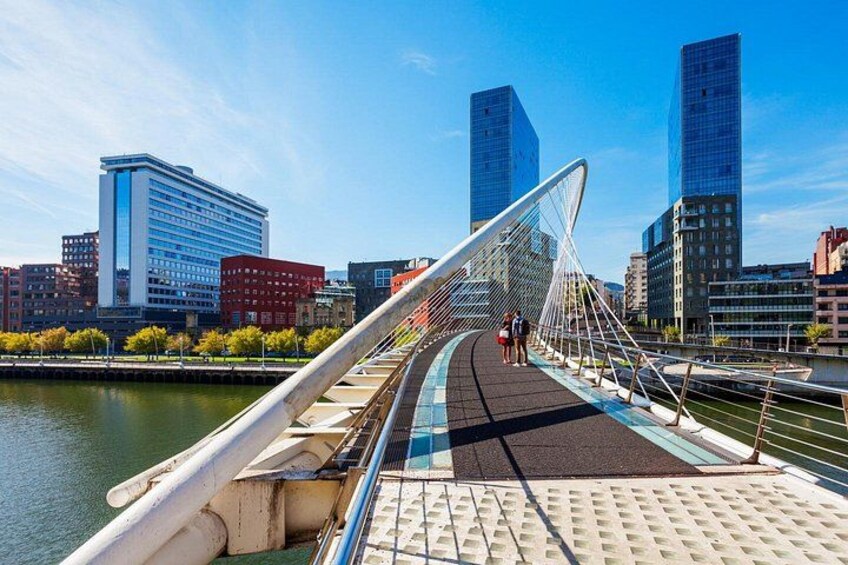 Eine Fußgängerbrücke mit Metallgeländer, ein Fluss und moderne Gebäude im Hintergrund. 2