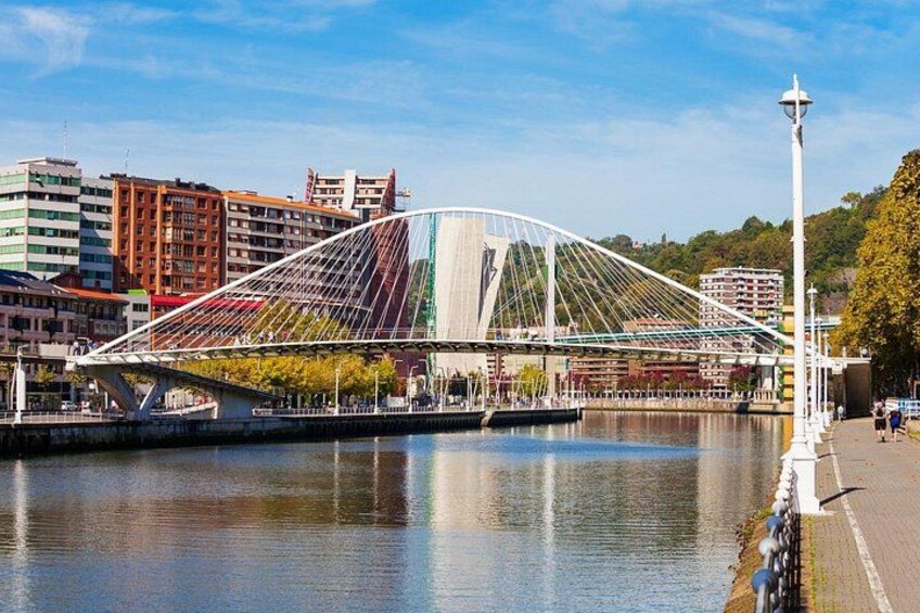Eine moderne Brücke mit Stahl- und Glasbauwerkskonstruktion überspannt einen Fluss, umgeben von Stadtgebäuden und wolkenlosem Himmel. 5