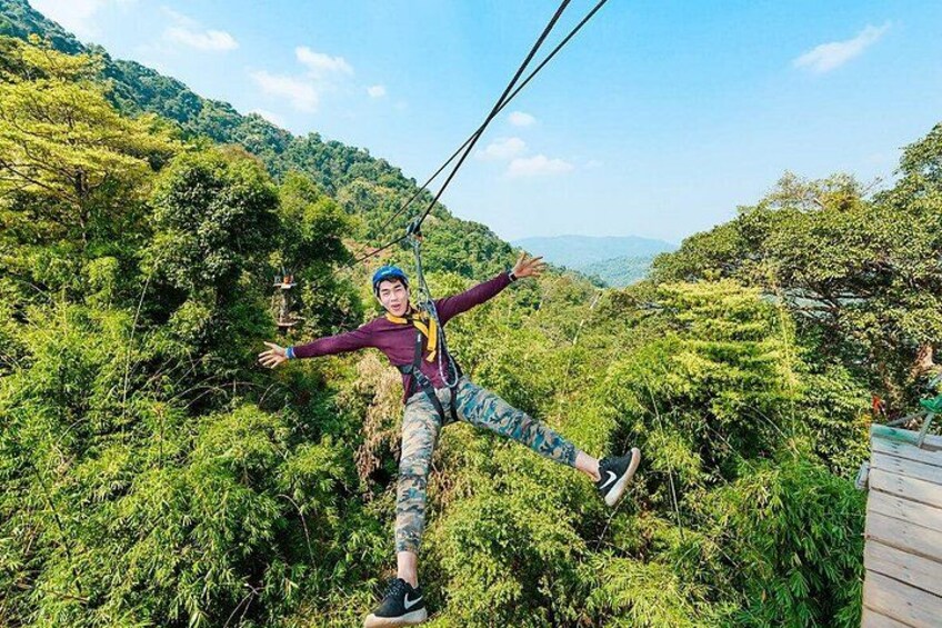 En person suser afsted i et zipline-anlæg gennem et skovområde med frodig vegetation og bakker i baggrunden. 1