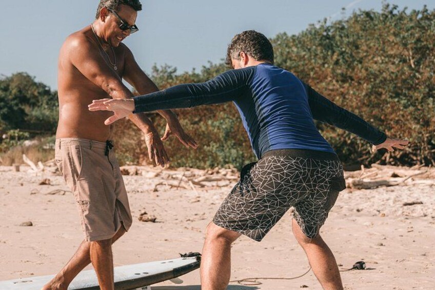 Two people on a sandy beach, one in swim trunks and the other in a long-sleeve shirt and patterned shorts, with a surfboard in the background. 4