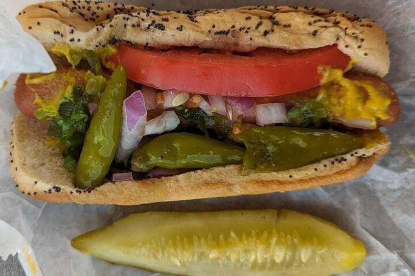 A hot dog with various toppings including tomatoes, onions, and pickles, and a pickle on the side. 2