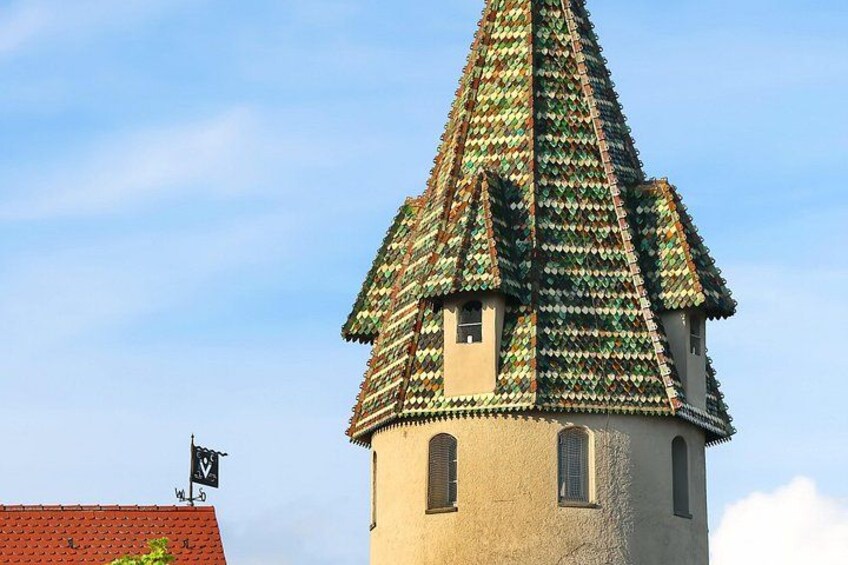 Et tårn med et kegleformet tag dækket af farverige fliser, en klar blå himmel og en bygning med rødt tag i forgrunden. 3