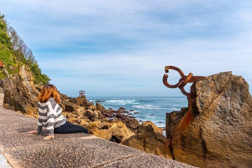 A person sitting on a rocky ledge overlooking the ocean, with a rusted metal sculpture in the foreground. 2