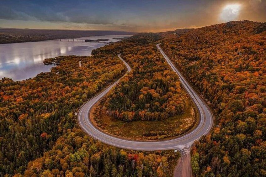 A winding road through a forested area with autumn foliage, a lake, and a bridge in the distance. 1