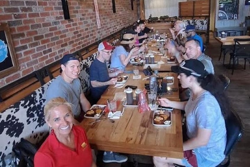 A group of people seated around a long wooden table in a restaurant with a brick wall and cow print booth seating. 2