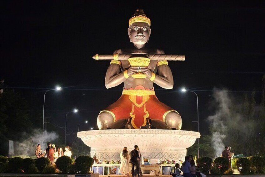 A large illuminated statue of a figure holding a weapon, surrounded by people and a stage setup. 1