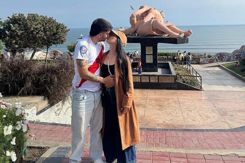 A couple embracing in a public square with a sculpture and a body of water in the background. 1