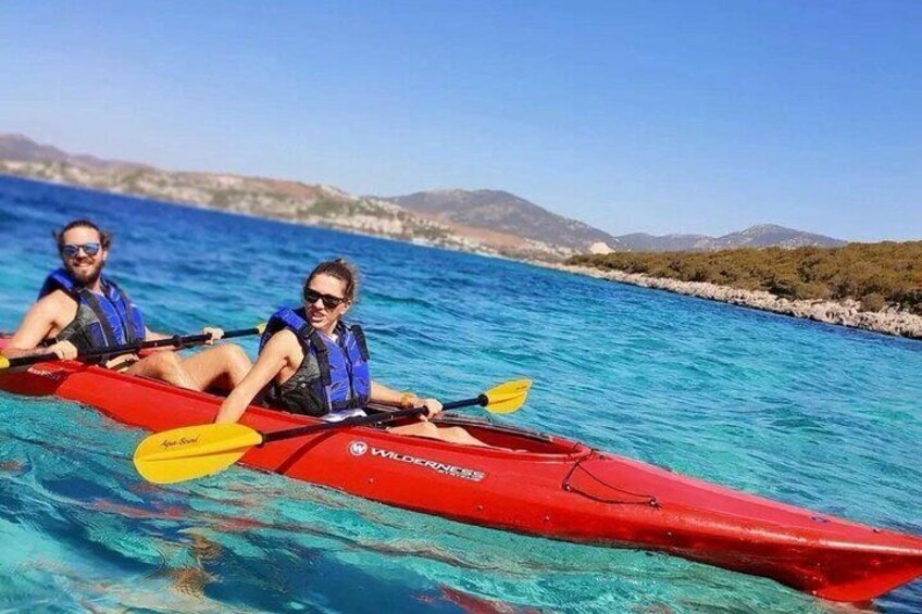 Two people kayaking on a clear blue sea with rocky coastline and hills in the background. 1