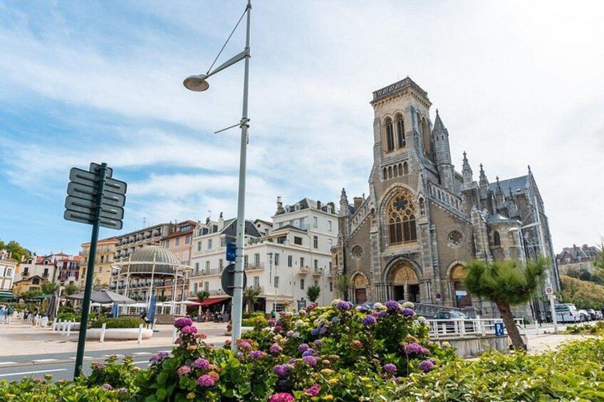 A historic church with a tall bell tower, surrounded by urban buildings and flowering bushes. 5