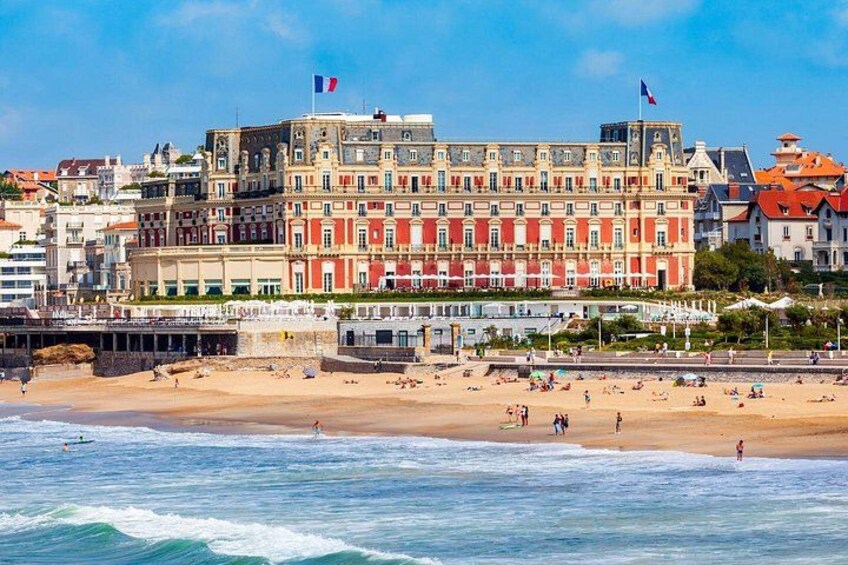 A beach with people, a large building with flags, and a pier. 2