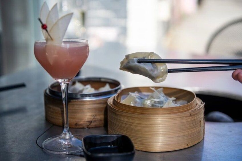 A drink in a glass, chopsticks holding dumplings, and a bamboo steamer with food. 1