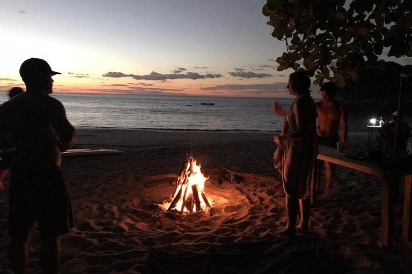 Une scène de plage avec un feu de camp, des personnes rassemblées autour, et l’océan en arrière-plan. 1
