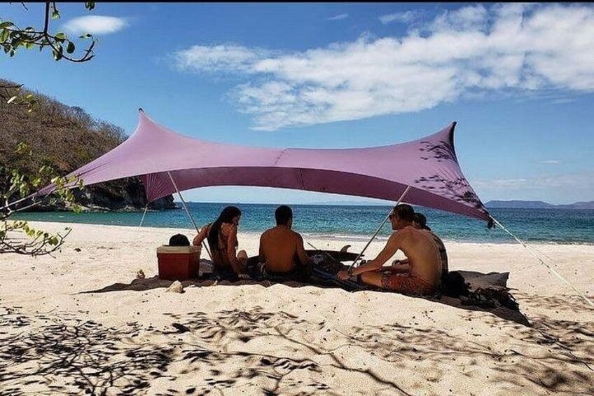 Trois personnes sont assises sur une plage, à l’ombre d’un parasol violet. En arrière-plan, on aperçoit un ciel dégagé et une mer calme. 4