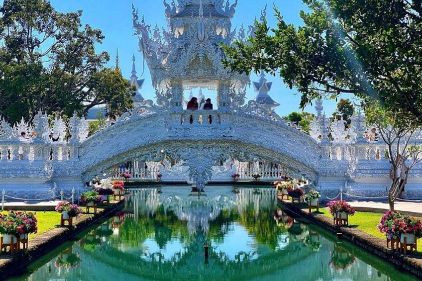 A white ornate temple with a reflection in the water, surrounded by trees and flowers. 3