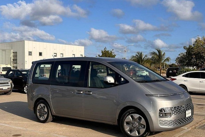 A silver minivan parked in a lot with a white building and palm trees in the background. 2