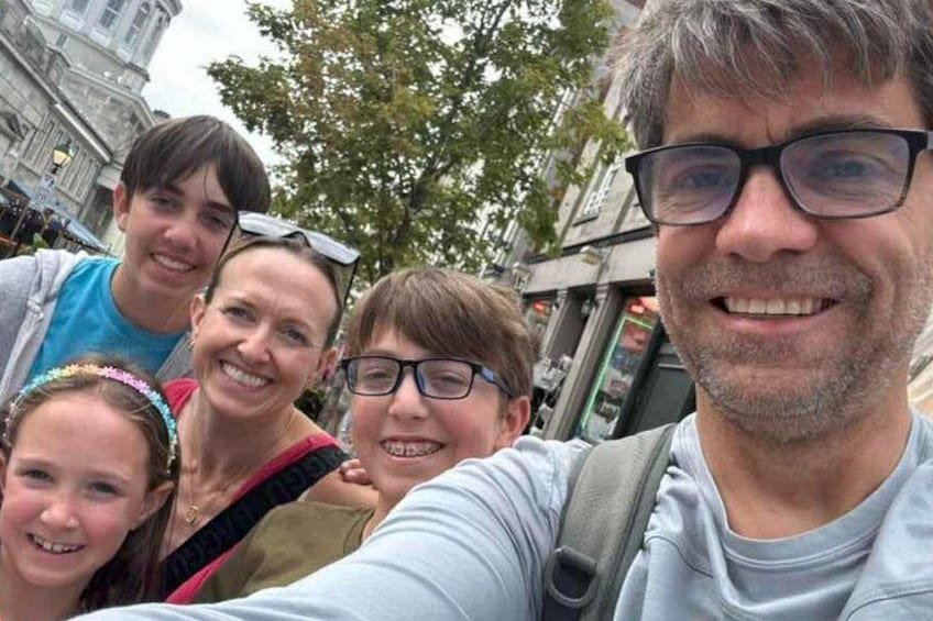 En grupp människor som tar en selfie i en stadsmiljö, med byggnader och träd i bakgrunden. 2