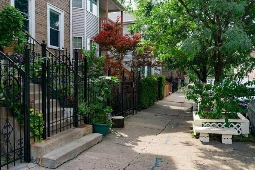 Une rue résidentielle bordée d’une clôture en métal noir, avec un banc blanc et divers pots de plantes. 4