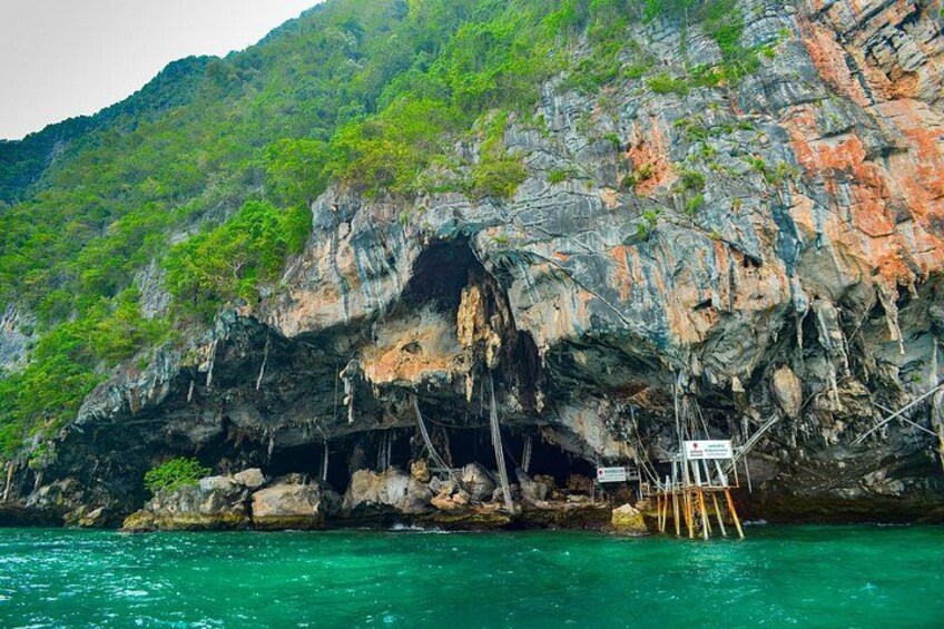 A rocky cliff with a cave entrance, a wooden platform, and clear turquoise water. 2