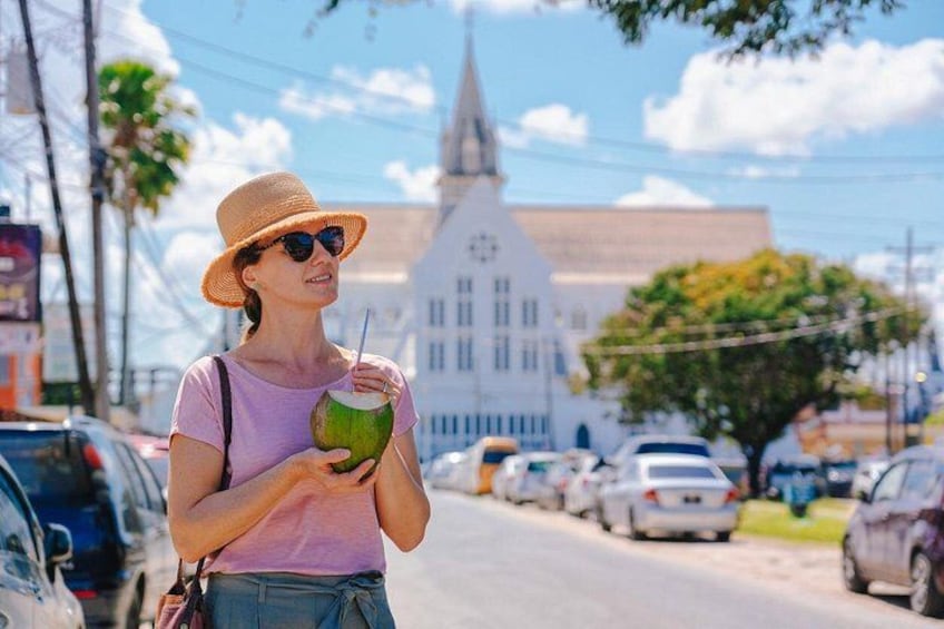 A person holding a coconut drink, standing on a street with parked cars and a church in the background. 5