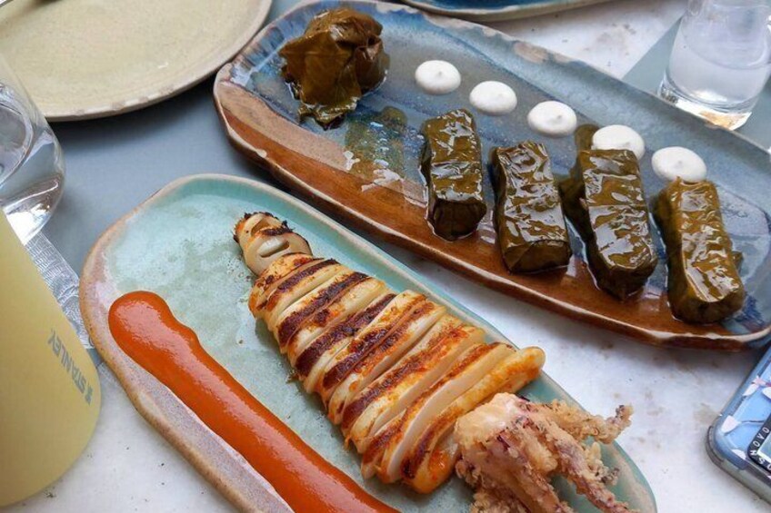 Un plato de hojas de parra rellenas y un guiso de carne a la parrilla, posiblemente cordero, sobre una mesa con un vaso de agua y un plato de panes planos. 2