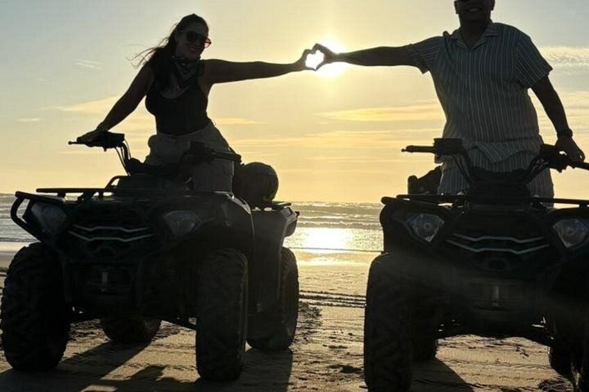 Due persone su quad ATV su una spiaggia, con il sole al tramonto sullo sfondo. 1