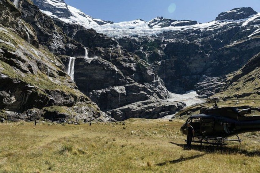 A helicopter parked in a mountainous area with a waterfall and snow-capped peaks in the background. 3