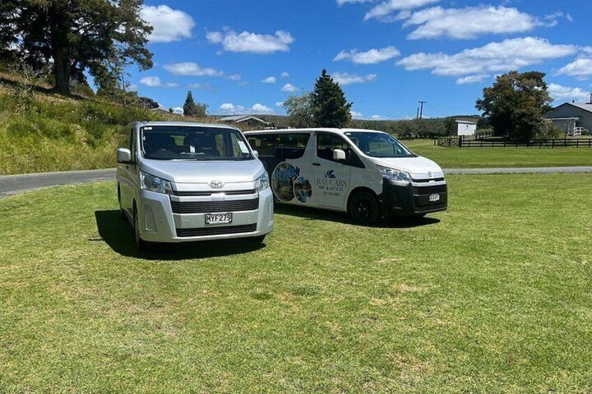 Two white vans parked on a grassy field with a clear blue sky and trees in the background. 2