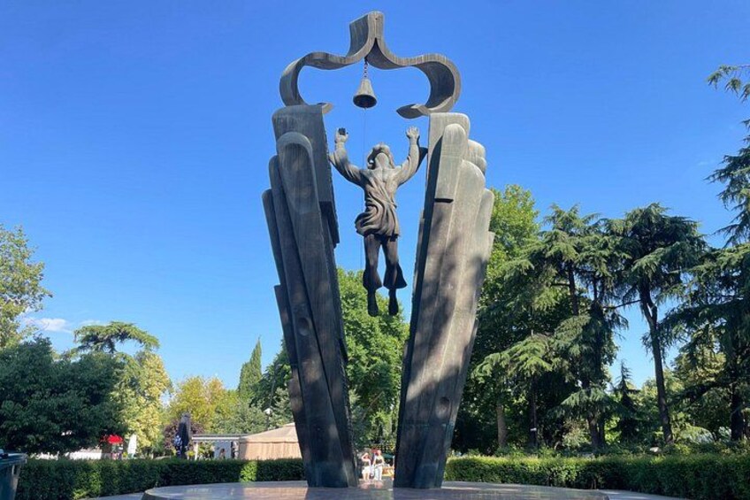 En skulptur som föreställer en person som håller i en klocka, placerad i en park med träd och en klar himmel. 1