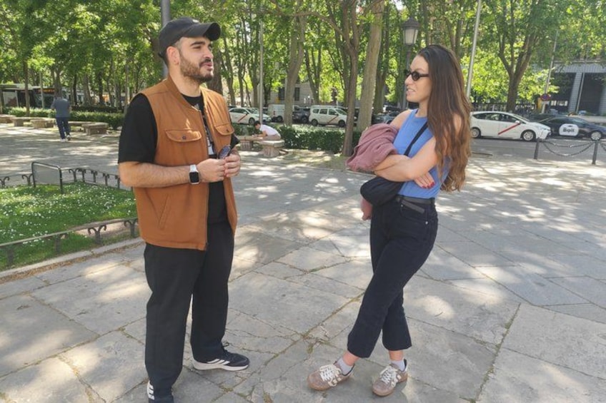 Two people standing on a paved area with trees and parked cars in the background. 4