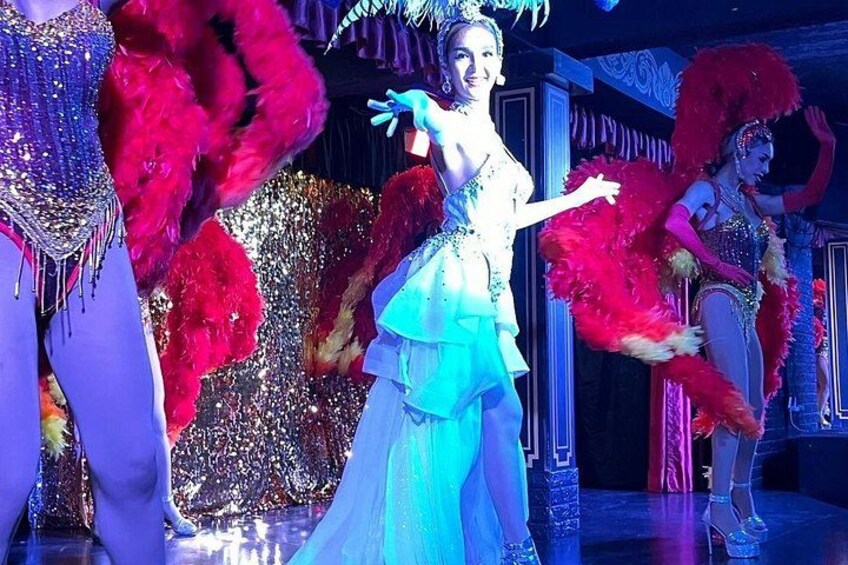 Un artista con un costume blu e bianco e una piuma di pavone in testa, che danza su un palco con uno sfondo scintillante. 5