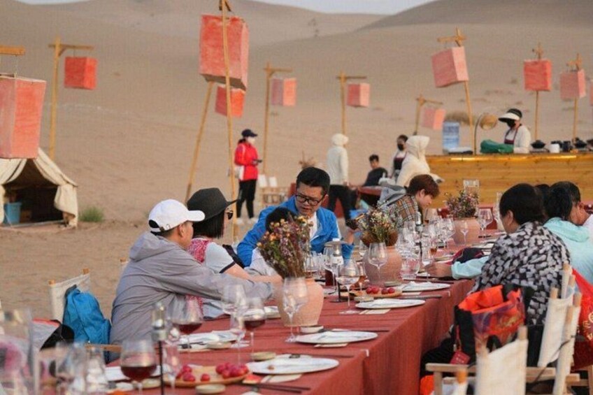 A group of people dining at a long table set up in a desert environment with tents in the background. 5