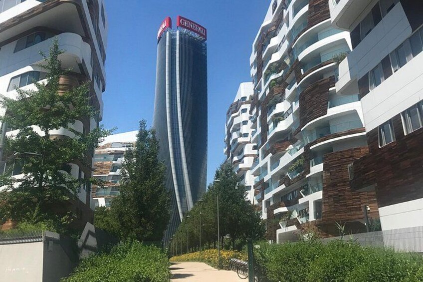 A modern urban area with high-rise buildings, a clear sky, and a pathway lined with greenery. 4