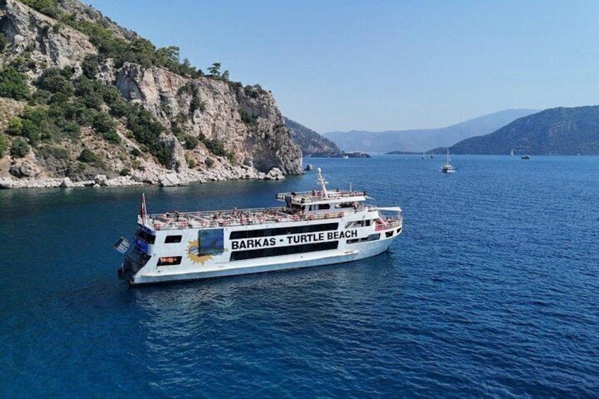 A ferry named 'Barkas - Turtle Beach' on a body of water with rocky cliffs and other boats in the distance. 1