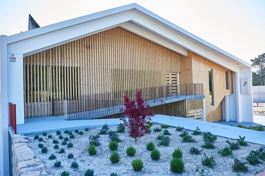 Un edificio moderno con techo inclinado, listones verticales de madera y un área ajardinada con plantas pequeñas y un árbol rojo. 2