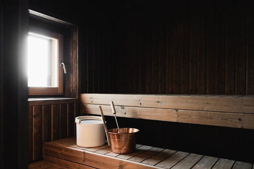 A wooden bench, a bucket, and a bucket of water in a cabin with a window. 1