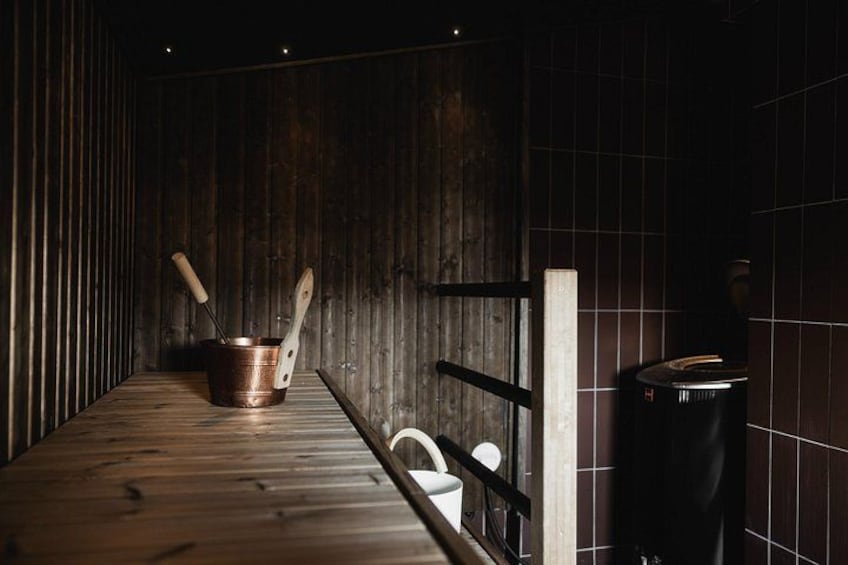 A wooden sauna bench, a copper bucket with a ladle, and a black heater in a dimly lit room with dark wooden walls. 3