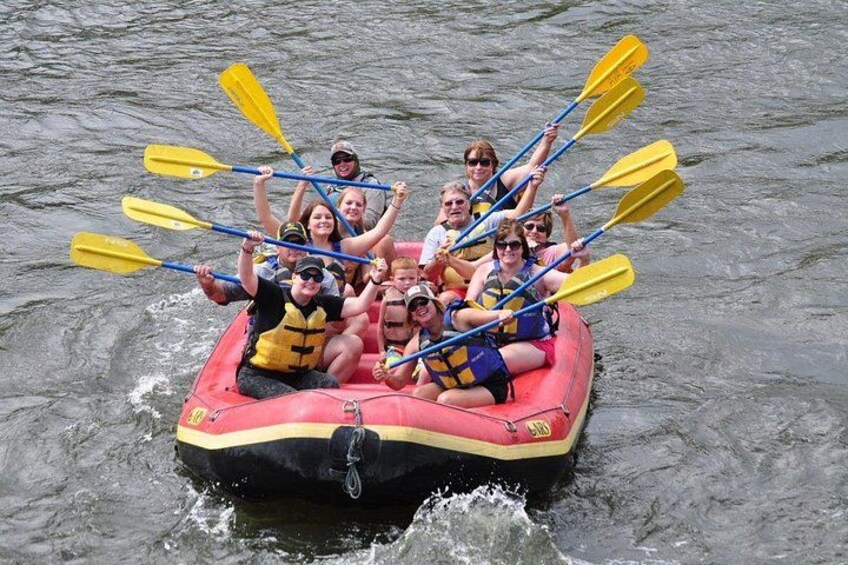 A group of people in a red inflatable raft on a body of water, paddling with yellow and blue paddles. 3