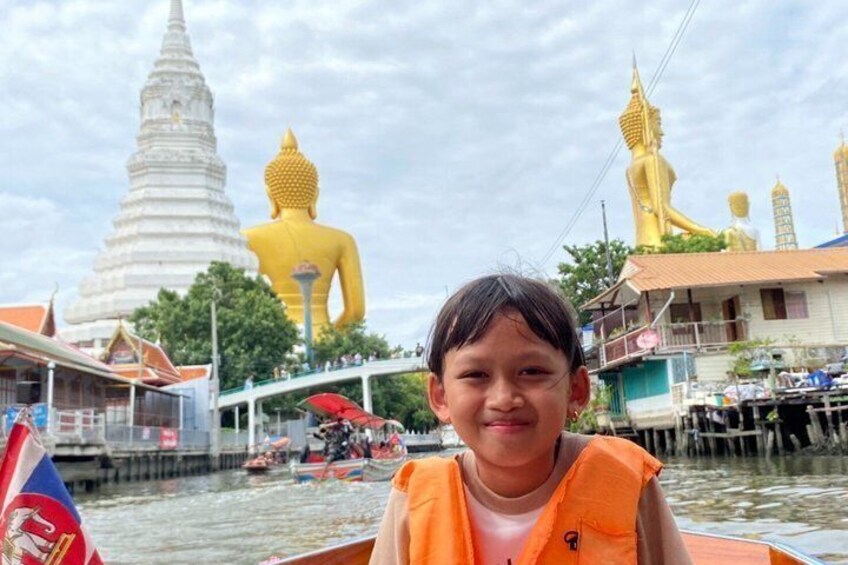 Eine Person in einem Boot vor großen, goldenen Buddha-Statuen und einer weißen Pagode im Hintergrund. 2