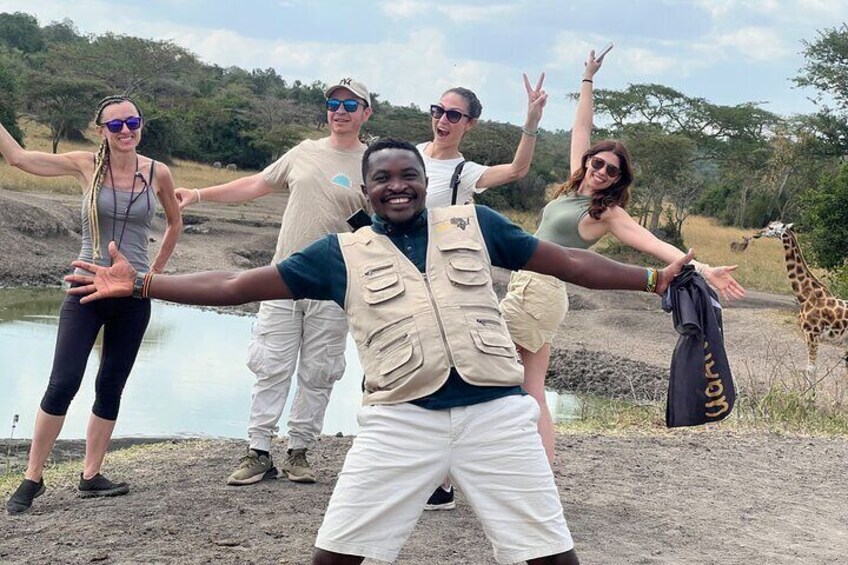 A group of people in safari attire posing by a water body with a giraffe in the background. 5