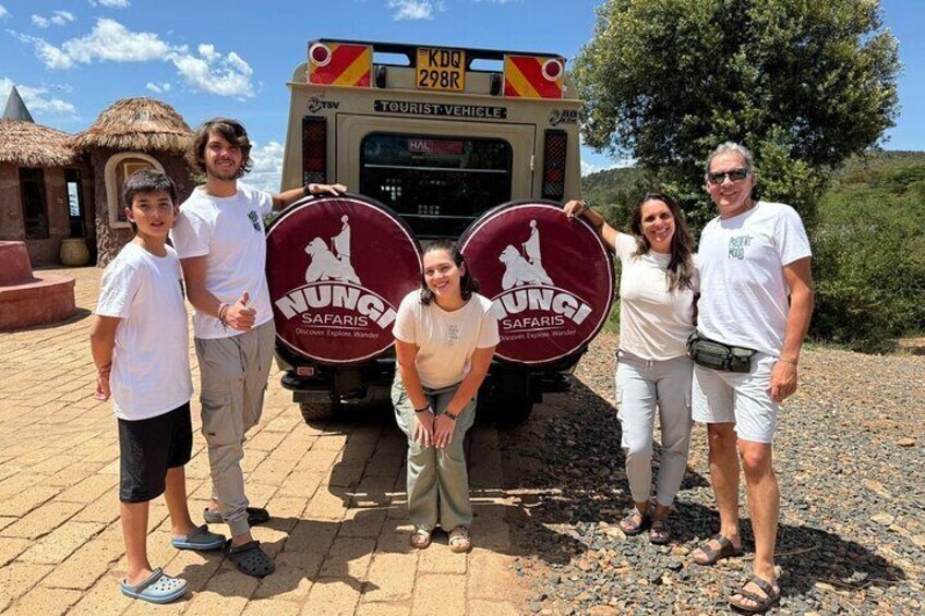 A group of people posing with a safari vehicle and two circular signs with 'HUNGRY SAFARIS' text. 1