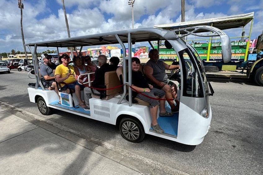 A small white vehicle with passengers, parked on a street with a colorful building in the background. 3