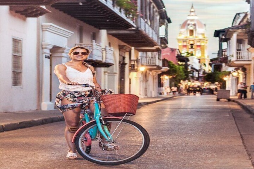 Iemand staat naast een turquoise fiets met een mandje, op een straat met gebouwen en een toren op de achtergrond. 1